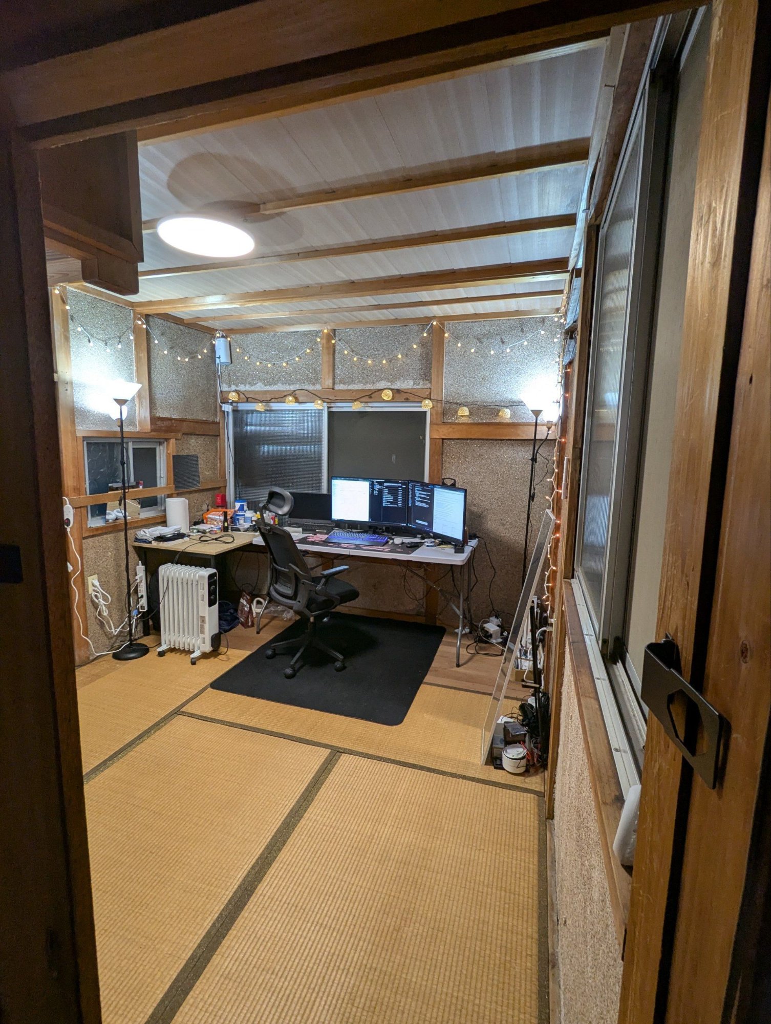 The Den at the Kaizen Dojo set up as a workspace: a multi-monitor desk on tatami matting under exposed beams and string lights.