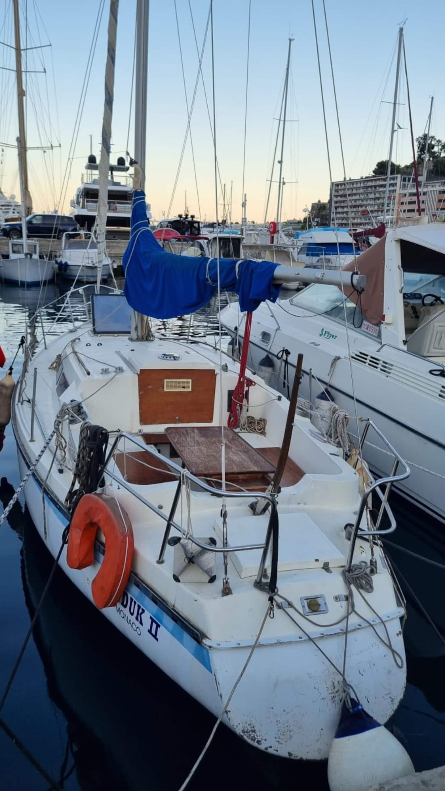 The foredeck of the Monaco sailboat looking forward over Port Hercule, with the bow, cleats and a blue sail cover in shot and other yachts moored alongside.